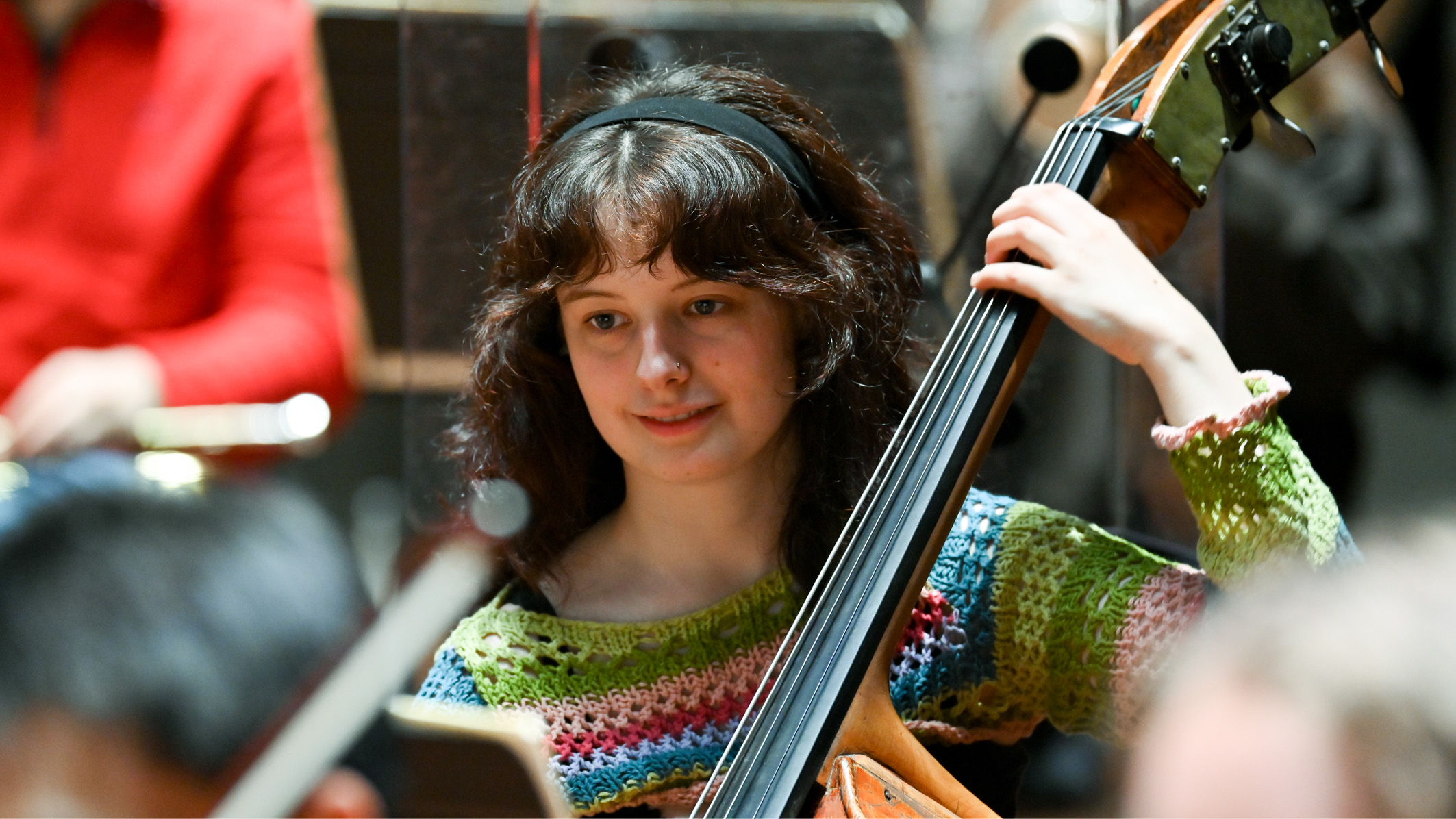 A female student playing a double bass in an orchestra rehearsal, smiling.